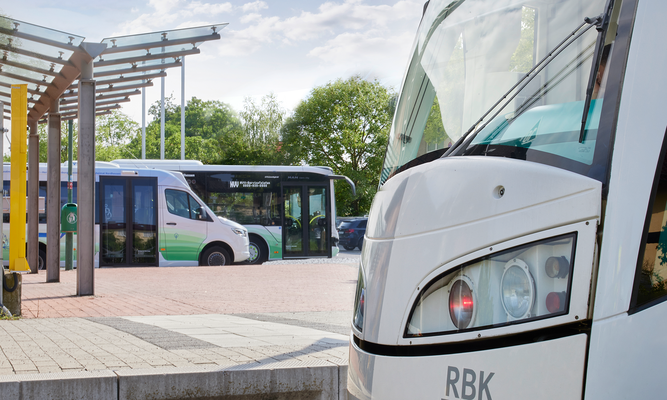 Nahaufnahme der Front eines weißen Fahrzeugs des öffentlichen Nahverkehrs. Im Hintergrund stehen ein weiterer Bus und ein Kleinbus an einer überdachten Haltestelle.