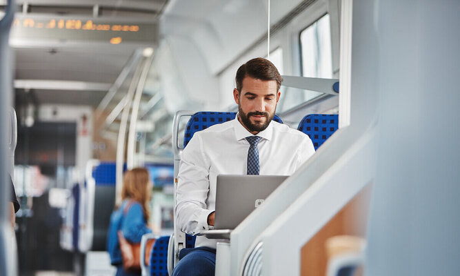 Ein Mann im Hemd und mit Krawatte sitzt in einem Zug auf einem blauen Sitz und arbeitet an einem Laptop. Im Hintergrund ist der Innenraum des Zuges mit weiteren Sitzplätzen und einer weiteren Person zu sehen; grafische Farbflächen in Blau und Grün sind eingeblendet.