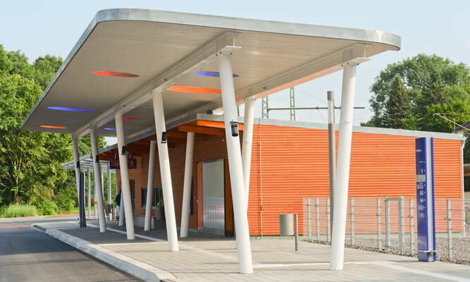 Moderne Bushaltestelle mit überdachtem Wartebereich und schräg stehenden weißen Stützen. Im Hintergrund befindet sich ein Gebäude mit orangefarbener Fassade; unter dem Dach sind farbige Lichtelemente sichtbar.