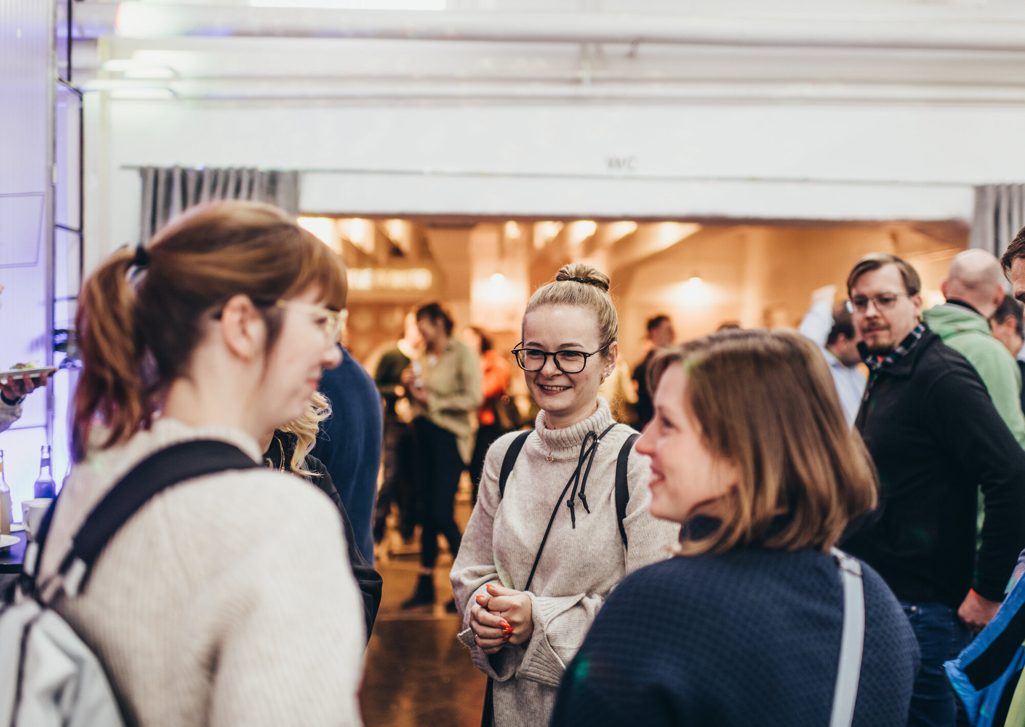 Mehrere Personen stehen bei einer Veranstaltung in einem Innenraum zusammen und unterhalten sich. Im Vordergrund sprechen drei Frauen miteinander, eine davon trägt eine Brille und lächelt. Im Hintergrund sind weitere Gäste zu sehen.