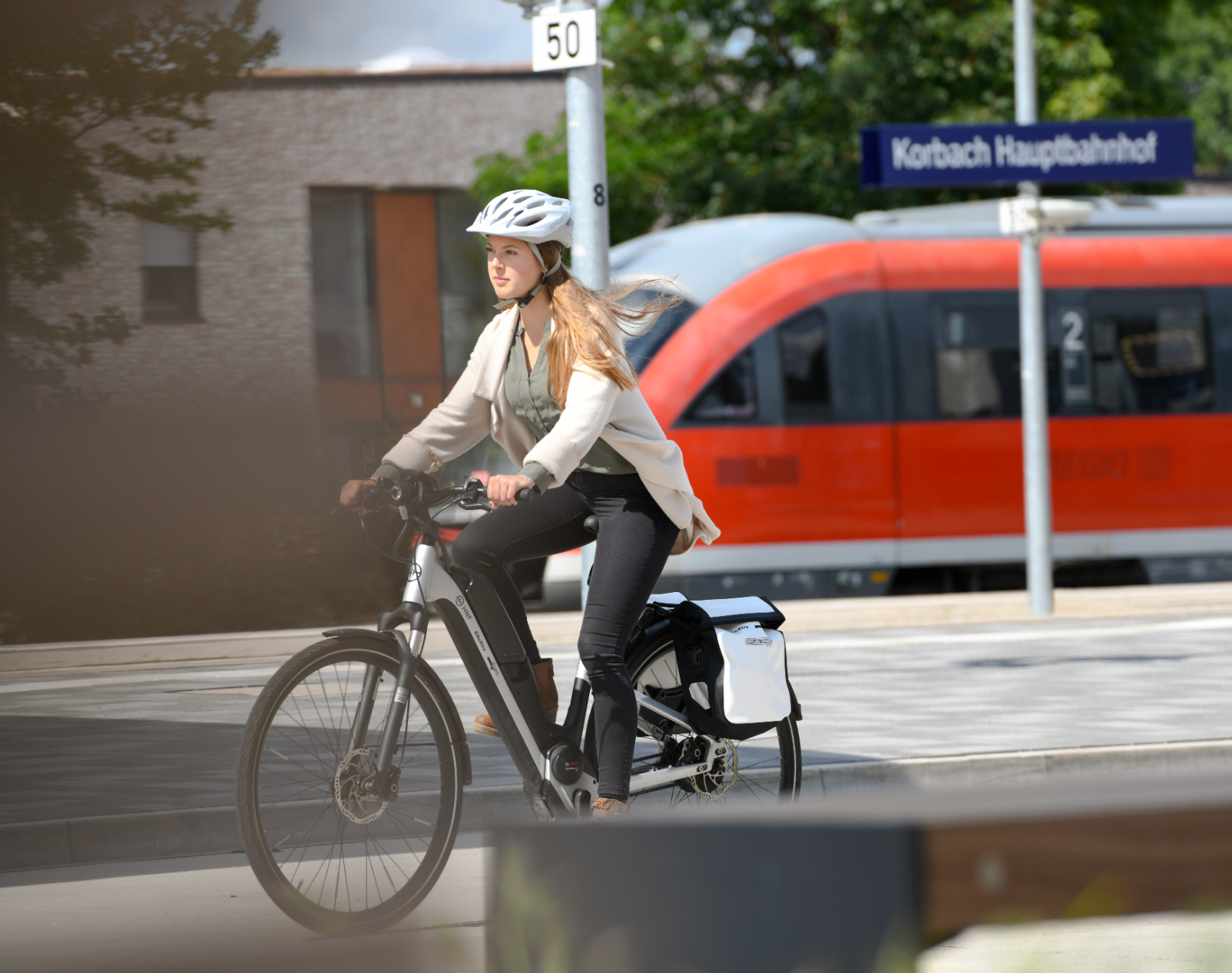 Eine Frau fährt am Bahnhof Fahrrad. Im Hintergrund ist eine Bahn zu erkennen.