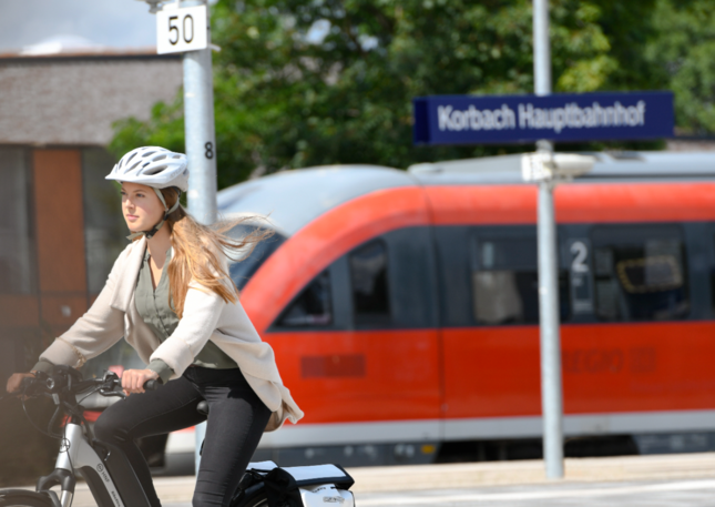 Eine Frau mit Fahrradhelm fährt mit einem E-Bike an einem Bahnhof entlang. Im Hintergrund steht ein roter Zug; ein Schild mit der Aufschrift „Korbach Hauptbahnhof“ ist zu sehen.